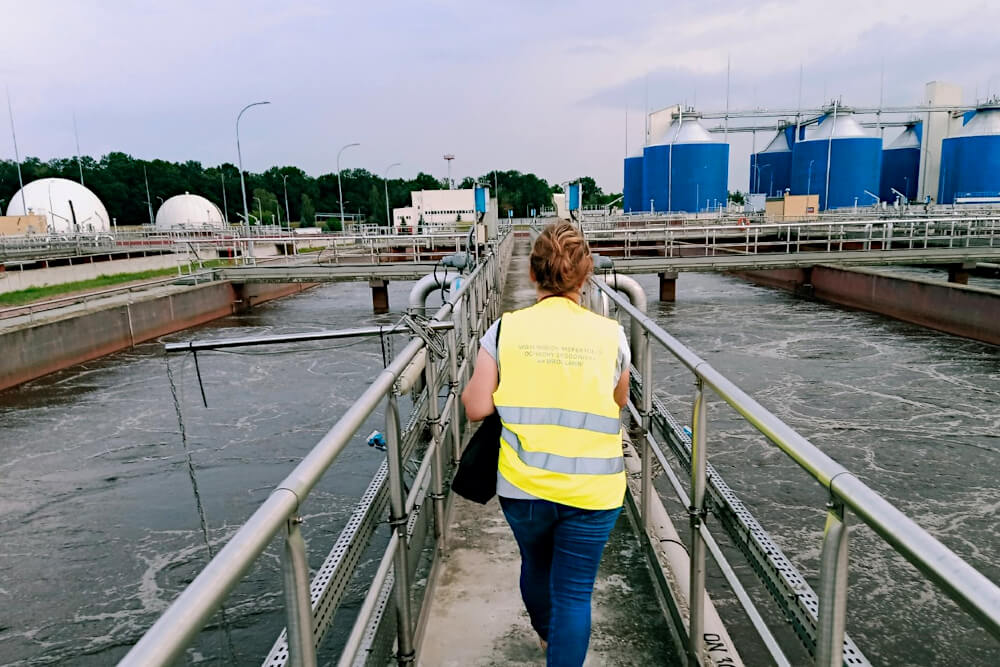 osoba stojąca tyłem z żółtej kamizelce z napisem Wojewódzki inspektorat Ochrony Środowiska we Wrocławiu, osoba idzie mostkiem, wokół mostku widoczna jest woda