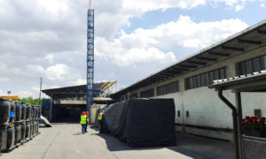 widoczne zabudowania magazynowe, w oddali wysoki komin, inspektorzy w kamizelkach i kaskach zaglądają pod plandekę okrywająca składowisko