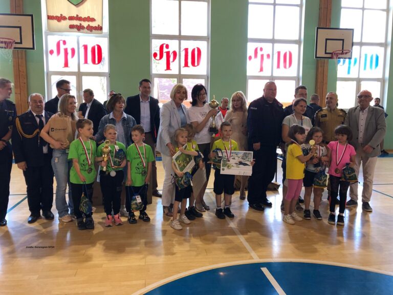 Widoczna sala sportowa, na której w szeregu na pierwszym planie stoją małe dzieci, trzymające w rękach dyplomy i puchary, za dziećmi w szeregu stoją osoby dorosłe, niektóre z nich są ubrane w mundury