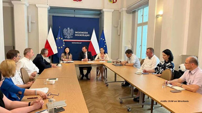 widoczne 2 długie stoły, ustawione równolegle przy którym siedzą różne osoby, na stole leżą dokumenty. Na głównej ścianie jest napis Dolnośląski Urząd Wojewódzki, po bokach dwie biało-czerwone flagi