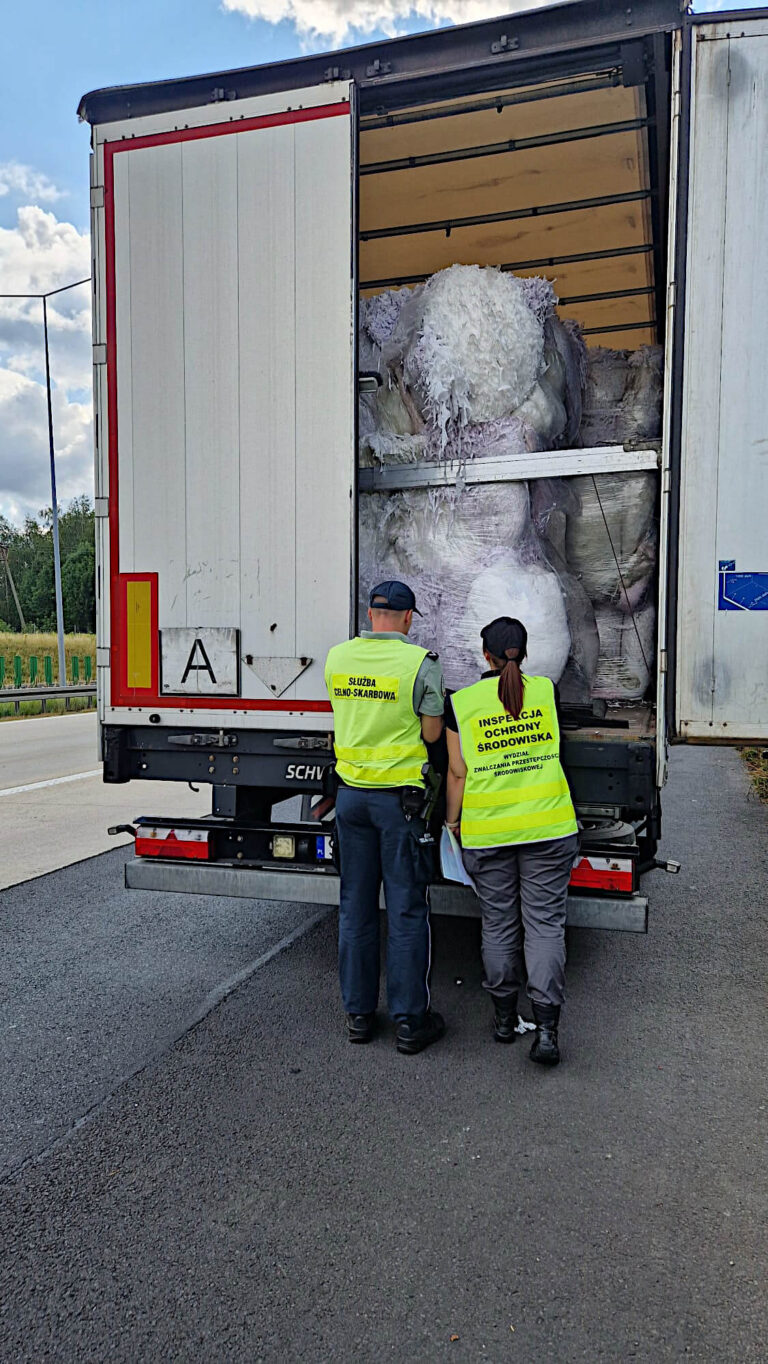 Widoczny tył naczepy samochodu ciężarowego, prawe tylne drzwi naczepy są otwarte i wewnątrz widoczne są wypełnione worki. Przy naczepie stoją dwie osoby, są odwrócone tyłem, ubrane w żółte kamizelki, jedna kamizelka ma napis „Inspekcja Ochrony Środowiska”, a druga „Służba Celno Skarbowa”.