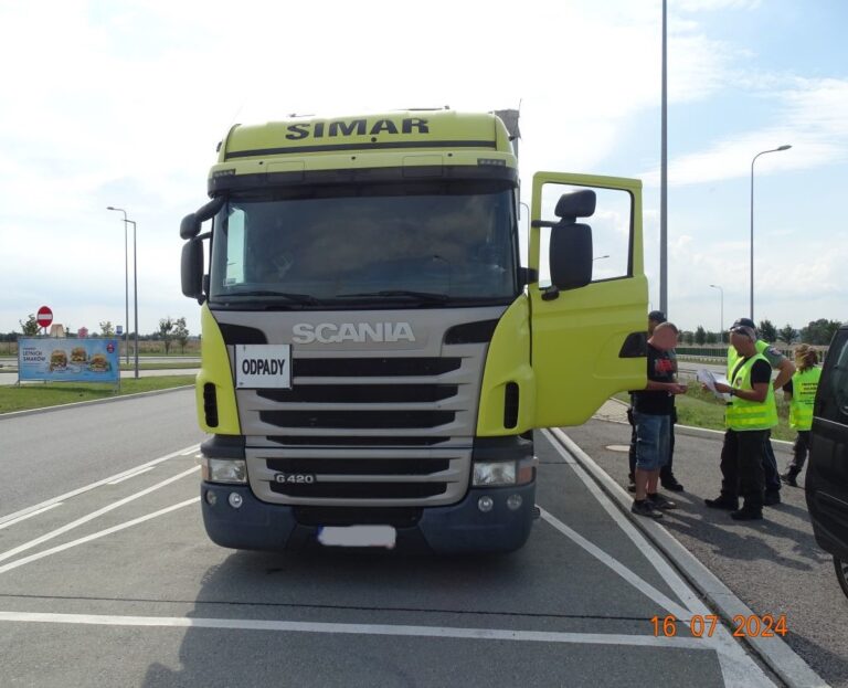 widoczny duży ciężarowy samochód "Scania" z drukowanym napisem "Odpady" po lewej stronie. Obok samochodu stoi kilka osób w żółtych kamizelkach