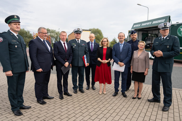 Ośmiu mężczyzn i dwie kobiety ustawione przodem do zdjęcia. Trzech z ośmiu mężczyzn ma na głowach garnizonowe czapki policyjne z godłem polskim. Meżczyzni ubrani są w granatowe garnitury oraz krawaty. Jedna z kobiet ma na nosie czerwoną sukienkę i czarną rozpiętą marynarkę. Druga kobieta jest ubrana w jasną koszulę i czarną długą spódnicę. W tle widoczny jest pojazd z napisem "Inspekcja transportu drogowego". Osoby znajdują się na parkingu samochodowym.