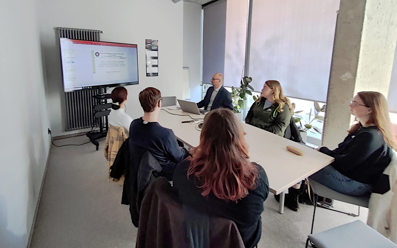 W małej sali konferencyjnej przy podłużnym drewniany stole siedzi 6 osób, w tym jeden prowadzący. Prowadzący to mężczyzna w średnim wieku siedzący najbliższej ekranu na którym wyświetlany jest slajd. Przy stole obok prowadzącego od jego lewej strony siedzą dwie młode kobiety a po przeciwnej stronie po środku siedzi młody mężczyzna a pomiędzy nim dwie młode kobiety. Wszyscy są studentami, którzy patrzą w stronę wyświetlanej prezentacji.