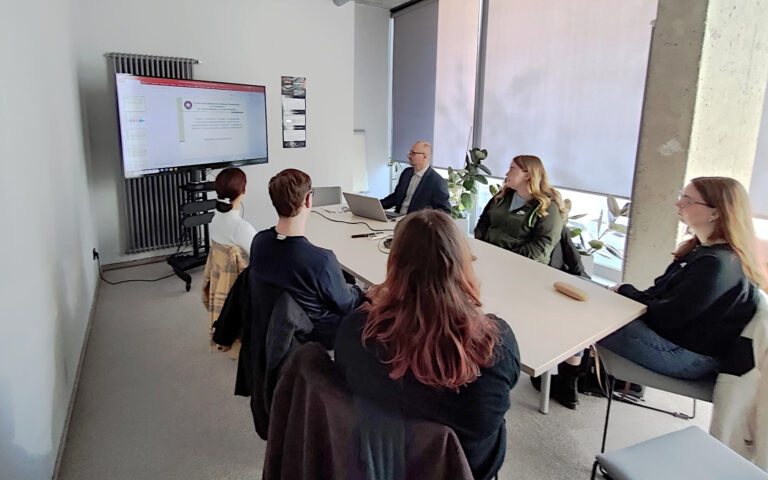 W małej sali konferencyjnej przy podłużnym drewniany stole siedzi 6 osób, w tym jeden prowadzący. Prowadzący to mężczyzna w średnim wieku siedzący najbliższej ekranu na którym wyświetlany jest slajd. Przy stole obok prowadzącego od jego lewej strony siedzą dwie młode kobiety a po przeciwnej stronie po środku siedzi młody mężczyzna a pomiędzy nim dwie młode kobiety. Wszyscy są studentami, którzy patrzą w stronę wyświetlanej prezentacji.