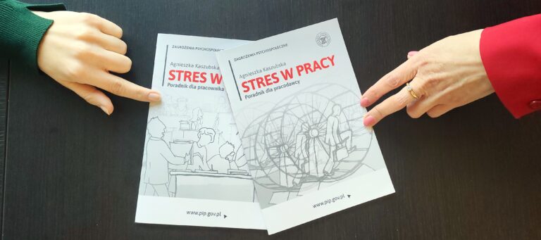 Na zdjęciu, w centralnym punkcie znajdują się dwa podręczniki, jeden nałożony na drugim. Są koloru białego. Napisane jest na nich czerwonym kolorem, drukowanymi literami "Stres w pracy". Egzemplarze są dedykowane zarówno dla pracowników jak i dla pracodawcy. Na podręczniki wskazują palce z dwóch osób, które są naprzeciwko siebie.