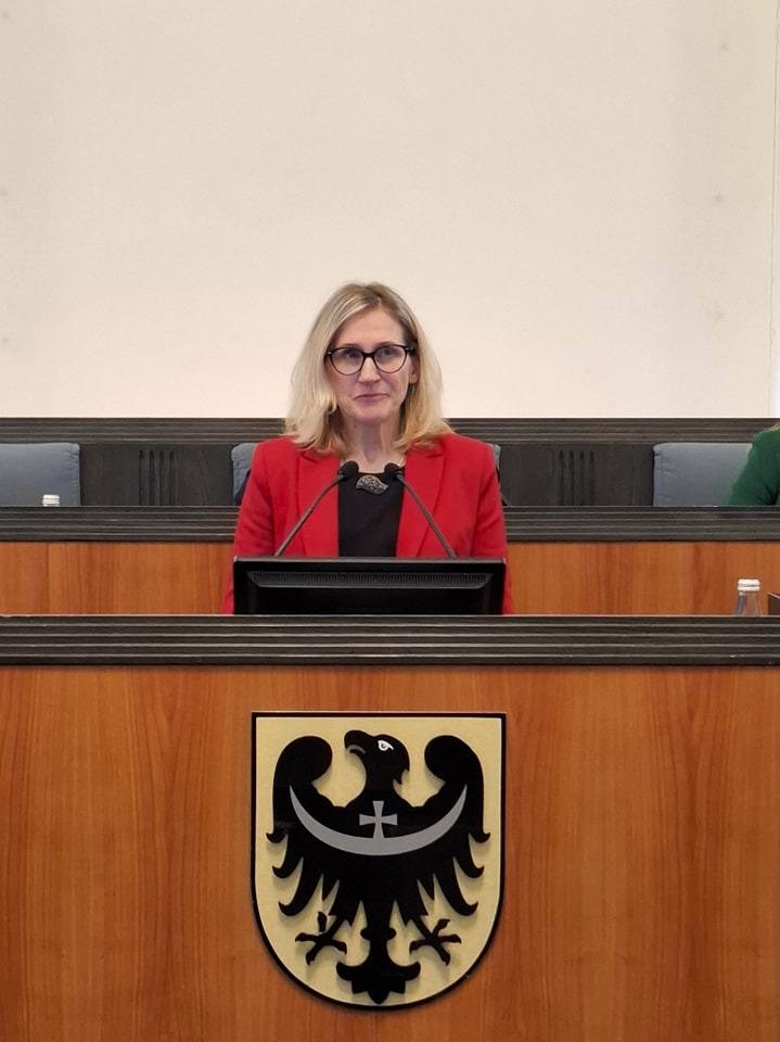 Przy mównicy znajduje się kobieta ubrana w czerwoną marynarkę. Skierowana jest do widowni. Z chwilą zrobienia zdjęcia przemawia. Na mównicy znajduje się herb województwa dolnośląskiego - w polu złotym orzeł czarny; przez tułów i skrzydła orła przepaska sierpowa srebrna, na niej pośrodku takiż krzyż