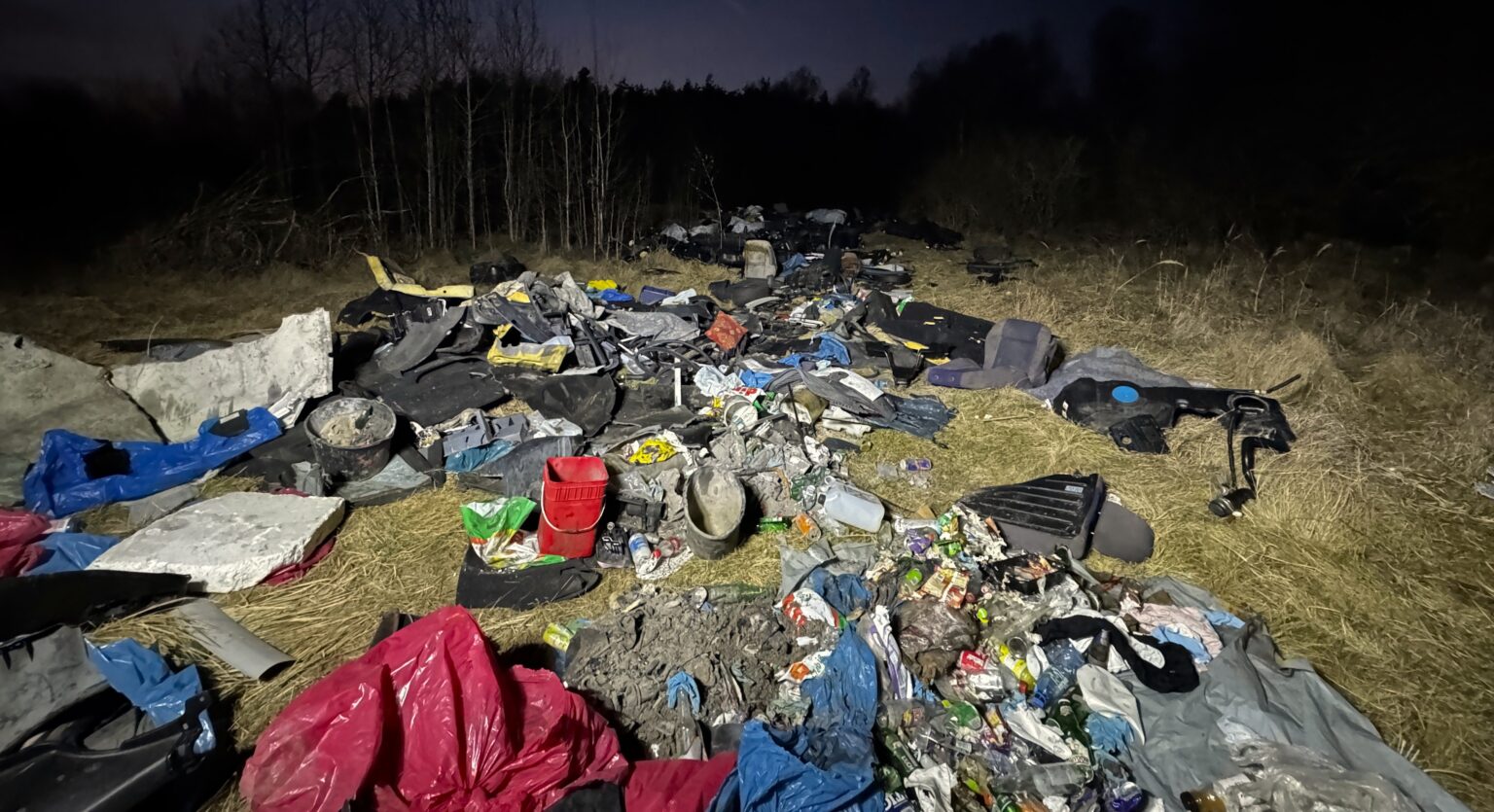Na zdjęciu widoczne są odpady zdeponowane na otwartej przestrzeni. Widoczne są odpady pochodzące z rozbiórki samochodów (opony, zbiorniki na paliwo, fotele) oraz ubrania, butelki, wiadra, opakowania po produktach chemicznych oraz gruz.