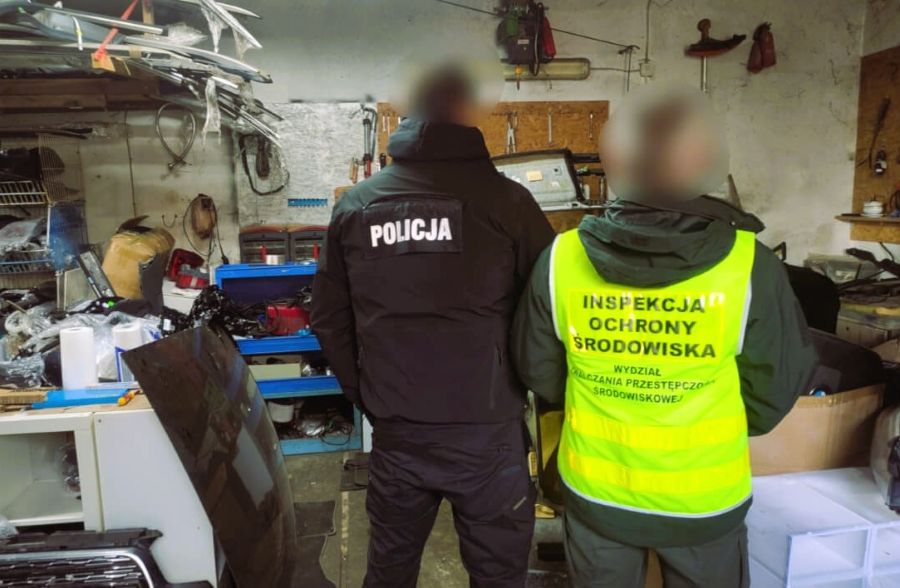 dwóch mezczyzn stojących tyłem do obiektywu. jeden z nich ma kamizelkę odblaskową z napisem Wojewódzki Inspektorat Ochrony Środowiska we Wrocławiu, Wydział Zwalczania Przestępczości Środowiskowej a drugi czarną kurtkę z napisem POLICJA. Męzczyzni stoją w pomieszczaniu, w którym znajdują się różnego rodzaju części samochodowe.