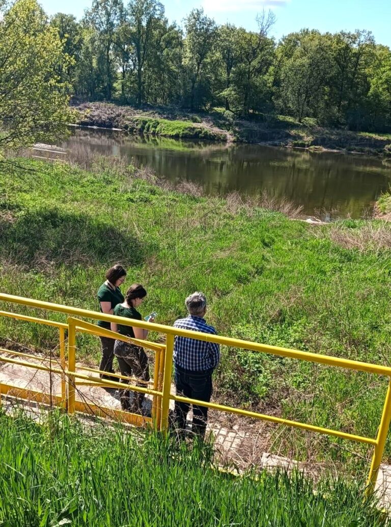 dwie kobiety i jeden mezczyzna stoją tyłem do obiektywu aparatu. znajdują na swieżym powietrzu na trawie, na wprost nich płynie rzeka odra. kobiety zapisują coś w swoich notatkach